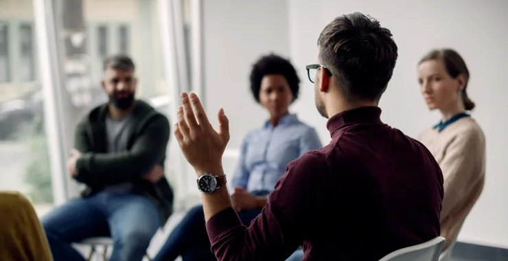 Rear view of man raising his hand to ask a question while attending group therapy.